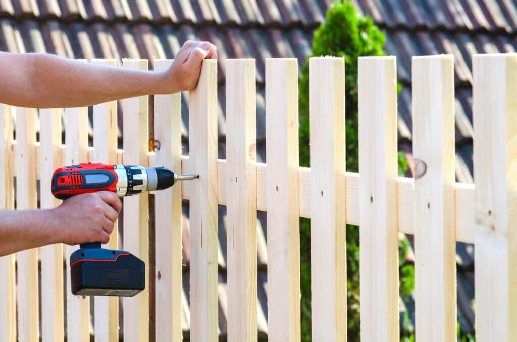 Un homme assemble une clôture en bois claire avec une perceuse sans fil, vissant une latte. Arrière-plan flou d'un toit et de verdure.